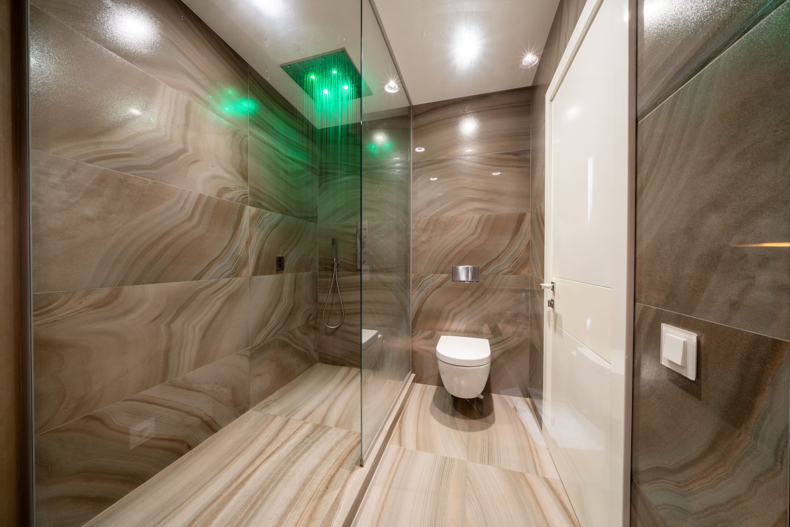salón de techos altos iluminado con grandes ventanales y leds baño en tonos marrones y luces integradas en el techo y ducha con iluminacion
